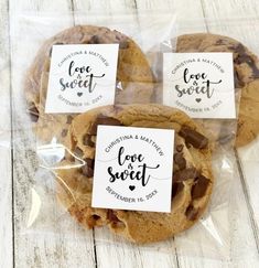 three chocolate chip cookies wrapped in plastic on a white wooden surface with love sweet stickers