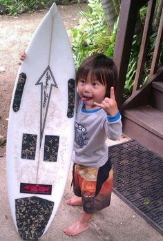 a little boy standing next to a surfboard giving the peace sign with his hand