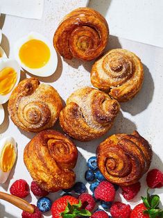 an assortment of pastries and berries on a white surface with eggs in the background