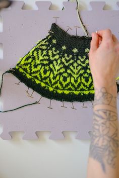 a hand is stitching a knitted hat on a piece of white paper with green and yellow designs