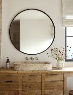 a bathroom with a round mirror above the sink and wooden cabinet below it, along with a vase filled with flowers