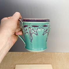 a hand holding a green coffee cup on top of a wooden table next to a white wall