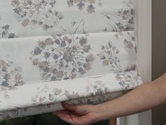 a hand is pulling the valance off of a flowered roman blind in front of a window