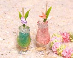 two small vases filled with different colored drinks on top of a sandy ground next to flowers