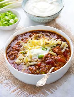 a white bowl filled with chili and cheese next to bowls of sour cream, celery and peas