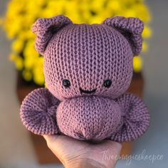 a hand holding a purple knitted teddy bear in front of some yellow and yellow flowers