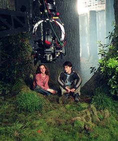 two people sitting on the ground in front of a tree with a camera attached to it