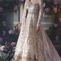 a woman standing in front of flowers wearing a white gown with gold embroidery on it