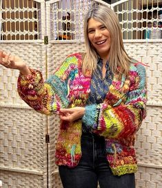 a woman standing in front of a white wall wearing a colorful cardigan sweater and jeans