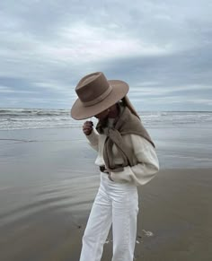 a woman standing on top of a beach next to the ocean wearing white pants and a brown hat