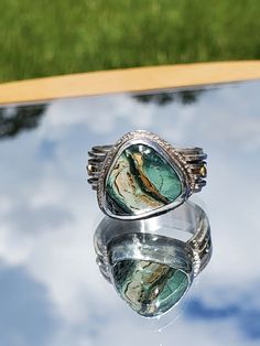 This ring features a 15 x 11mm Turquoise gemstone set in Sterling Silver and accented with two, 14kt gold balls.  The turquoise features bands of gold, cream, light green and dark green.  It is cut in a triangle shape and set at an angle to best compliment a woman's finger.  The band is 10 mm wide and features 4 individual bands of Sterling Silver to give it a multi-dimensional look.  The gold balls accent the gold in the turquoise. This is a one-of-a-kind ring that you will not see anywhere else. RETURN POLICY: I accept returns under the following conditions.  The jewelry must be returned in new condition.  Purchaser must contact me within 3 days of receipt of the jewelry.  The ring must be returned in the same jewelry box it was shipped in, and it must be returned vias USPS Priority Mail Green Turquoise Ring With Natural Stones, Bohemian Green Rings With Gemstone Accents, Green Chrysocolla Rings With Natural Stones, Artisan Green Chrysocolla Rings, Unique Turquoise Rings With Gemstone Accents, Unique Green Emerald Ring With Gemstone Accents, Unique Green Ring Jewelry, Unique May Birthstone Jewelry With Accent Stones, Green Chrysocolla Cabochon Jewelry