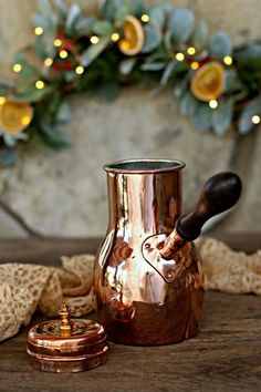 a copper pitcher sitting on top of a wooden table next to a christmas wreath with lights in the background