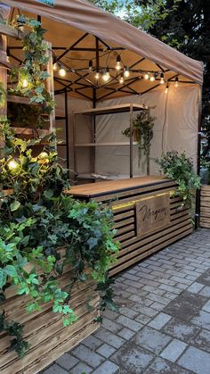 an outdoor food cart with plants growing on the outside and lights hanging from it's roof