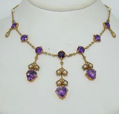 This necklace features fabulous deep purple amethysts. It is crafted from 14K yellow gold, natural amethysts and seed pearls. This necklace dates from the 1900s. It is presented in excellent condition with no repairs or damage. The necklace features three heart drops from the center of the necklace out. The drops encompass a heart cut amethyst as well aa seed pearls adorned on a gold leaf motif. The drops dangle from the body of the necklace. The body features prong set round cut amethysts, two Purple Sapphire Necklace, Victorian Purple Necklace For Anniversary, Antique Purple Necklace For Wedding, Body Features, Crystal Choker Necklace, Leaf Motif, Purple Sapphire, Gold Bead Necklace, Coral And Gold