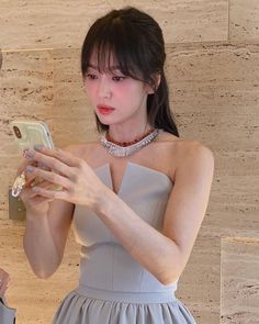 a woman in a gray dress is looking at her cell phone and holding a necklace