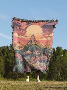 someone holding up a blanket in the middle of a field with mountains and clouds on it