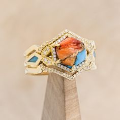 a gold ring with an orange and blue stone surrounded by small white diamonds on a wooden stand