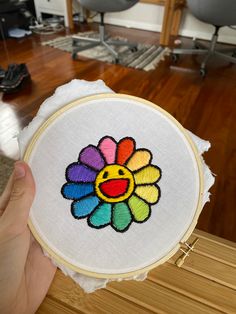 someone is holding up a colorful flower embroidery on a piece of fabric in front of a wooden table