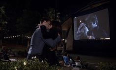 two people embracing each other in front of a movie screen at night with lights on