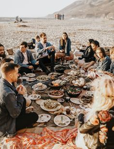 a group of people sitting around a table eating food on top of a sandy beach