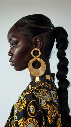 a woman with black hair wearing large gold hoop earrings and a patterned top, standing in front of a white wall