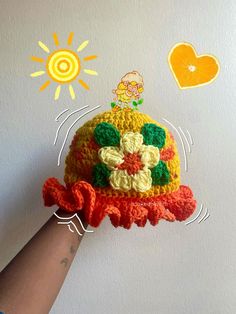 a hand holding a crocheted hat with an orange heart and sun above it