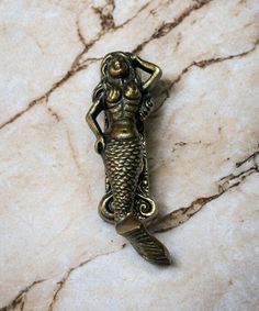 a gold colored mermaid brooch with a fish tail on it's back, sitting on a marble surface