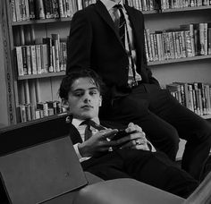 two young men sitting in front of bookshelves, one holding a game controller