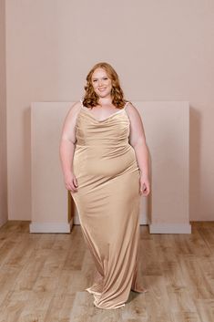 a woman in a tan dress standing on a wooden floor with her arms behind her back