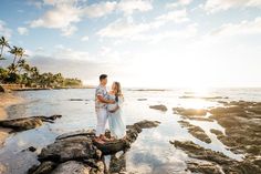 a man and woman standing on rocks near the ocean at sunset with palm trees in the background