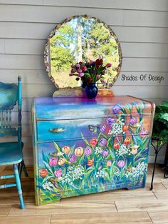 a painted dresser with flowers on it next to a blue chair and a round mirror