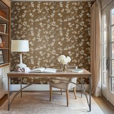 a desk with a lamp and flowers on it in front of a wallpapered room