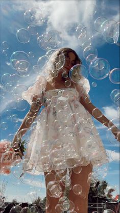 a woman in a white dress is surrounded by soap bubbles on a sunny day with blue sky and clouds behind her