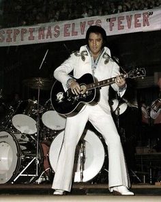 elvis presley performing on stage with his guitar and bandleader in the background at a concert