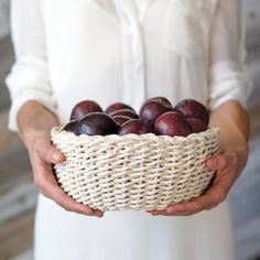 a woman is holding a woven basket with plums in it while wearing a white shirt