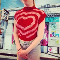 a woman standing in front of a building wearing a red knitted heart sweater and jeans