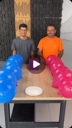 two men standing next to each other at a table with blue and pink balloons on it