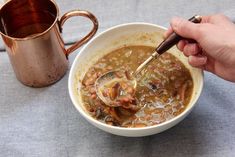 a person holding a ladle in a bowl of soup next to a copper mug