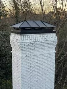 a white brick chimney with a black roof in front of some trees and shrubbery