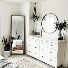 a bedroom with white furniture and plants on the dresser next to a large round mirror