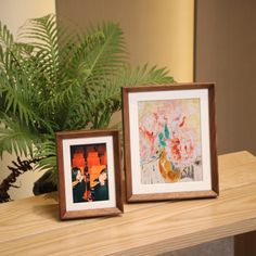 two framed pictures sitting on top of a wooden table next to a potted plant