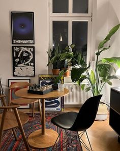 two chairs and a table in a room with plants on the windowsill, and posters above them