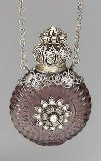 an old fashioned perfume bottle is sitting on a silver chain, with a flower in the center