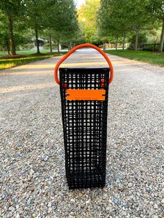 an orange and black trash can sitting on top of a gravel covered road next to trees