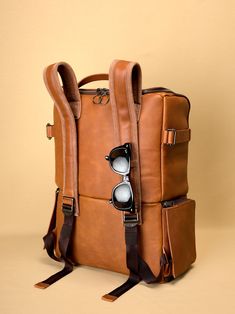 a brown leather backpack with sunglasses on the front and strap around the bottom, sitting against a tan background