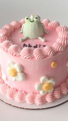 a pink cake decorated with white flowers and a frog sitting on it's top