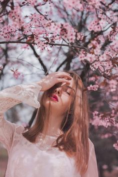 a woman standing under a tree with pink flowers on it's branches and her eyes closed