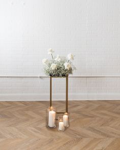 two vases filled with white flowers and lit candles on a wooden floor next to a brick wall