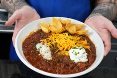 a person holding a bowl of chili with cheese and tortilla chips on the side