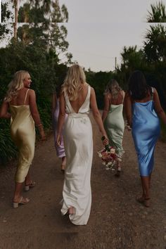 four women walking down a dirt road holding hands and looking at each other's back